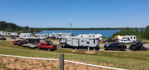 A scenic view of RVs and trucks parked by a lake on a sunny day, surrounded by greenery.