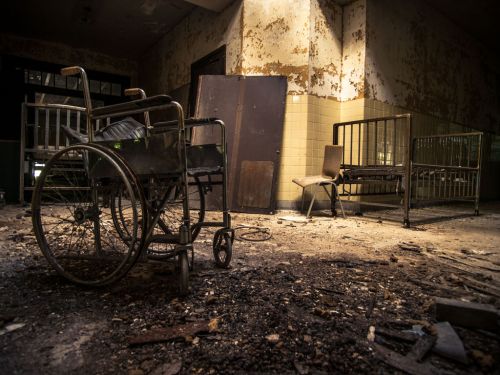 Abandoned room with old wheelchairs and hospital beds, surrounded by dust and debris in a decaying environment.