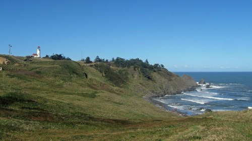A scenic coastal view featuring a lighthouse on a grassy hill overlooking the ocean and waves. Clear blue sky above.