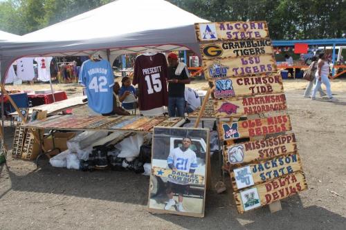 A vendor booth displaying sports jerseys and signs for various college teams at an outdoor market.