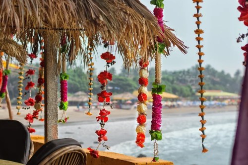 Colorful floral decorations hang from a thatched roof, overlooking a beach with umbrellas and gentle waves in the background.