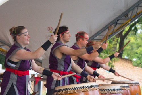 A group of four performers in traditional attire playing taiko drums under a tent, focused and energetic.