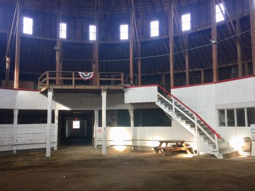 Interior of a round barn with wooden beams, a staircase, and a picnic table, illuminated by natural light.