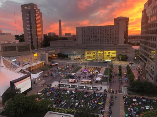 A vibrant sunset over a city plaza filled with people on blankets, food trucks, and a large screen for a movie.