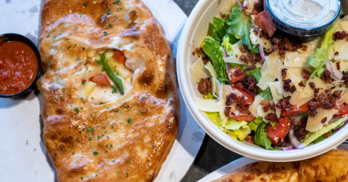 A golden-brown calzone next to a fresh salad topped with bacon, cheese, and a small container of dressing.