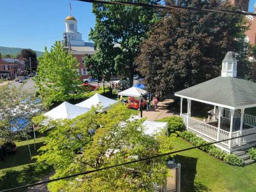 A sunny park scene with tents set up for an event, surrounded by trees and a gazebo, with a clock tower in the background.