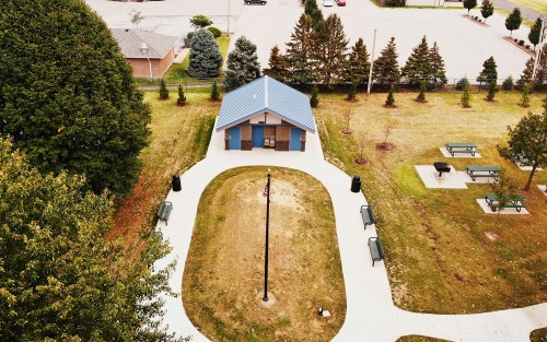 Aerial view of a park with a small building, picnic tables, and a flagpole surrounded by trees and grassy areas.