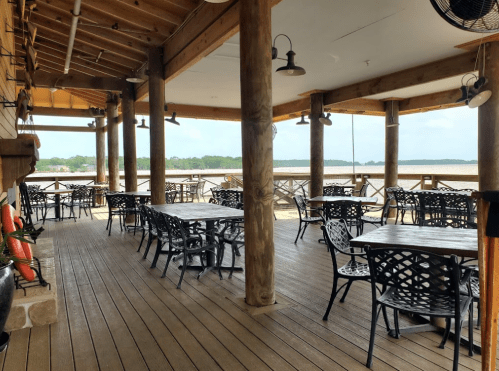 A spacious outdoor dining area with wooden floors, tables, and chairs overlooking a calm water view.