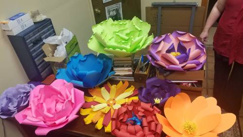 Colorful paper flowers in various sizes and shapes arranged on a table, with crafting supplies in the background.