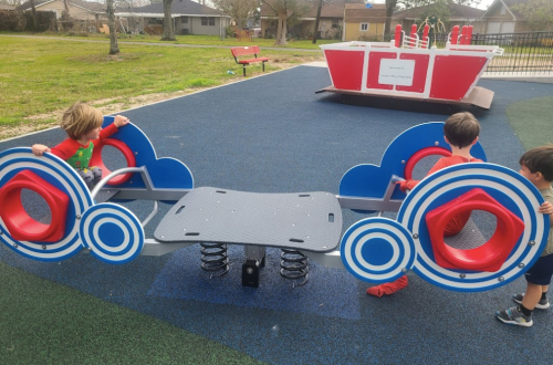 Three children play on a colorful seesaw at a playground, with a red and white play structure in the background.