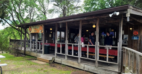 A rustic wooden restaurant with a porch, surrounded by trees, featuring outdoor seating and patrons inside.