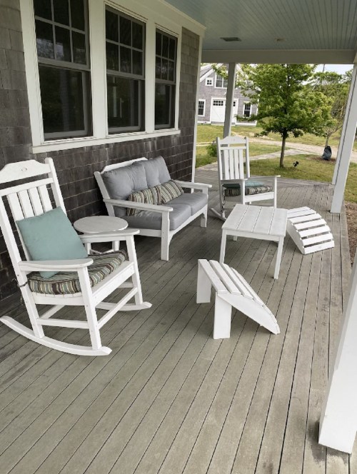 A cozy porch with white rocking chairs, a sofa, and a small table on a wooden deck surrounded by greenery.