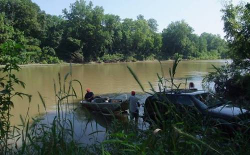 Two people in a small boat near a muddy riverbank, surrounded by lush greenery and trees.