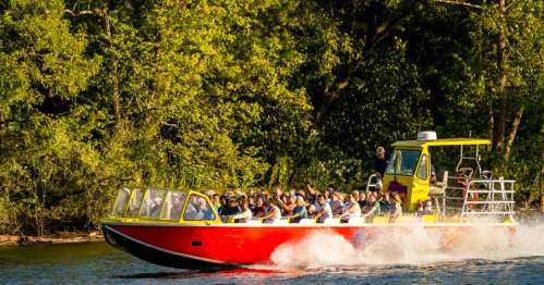 A bright red boat with passengers speeds across a river, surrounded by lush green trees.