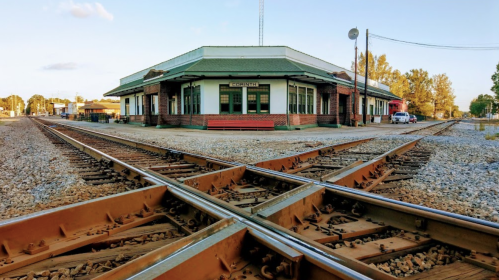 Historic train station with a green roof, surrounded by intersecting railway tracks and a clear blue sky.