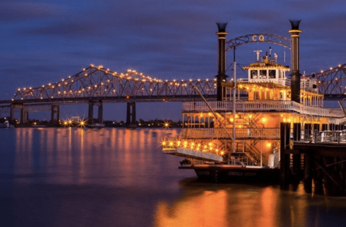 A brightly lit riverboat docked by a bridge at dusk, reflecting on the calm water.