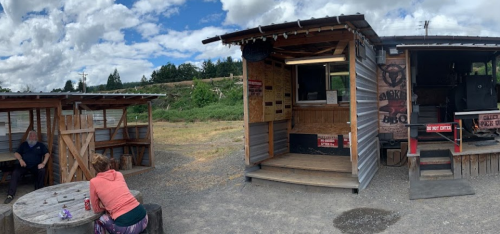 A rustic BBQ food truck with outdoor seating, surrounded by greenery and a partly cloudy sky.