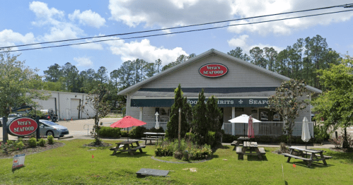 Exterior of Vera's Seafood restaurant with outdoor seating, umbrellas, and a sign, surrounded by greenery and blue sky.