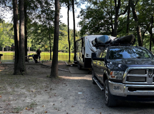 A truck parked near a camper by a serene lake, surrounded by trees, with a person at a picnic table in the background.