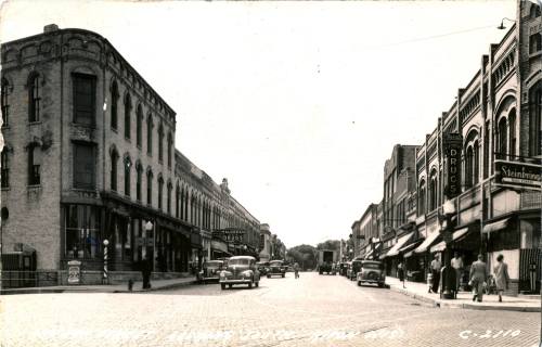 Historic black-and-white photo of a downtown street with vintage cars, shops, and pedestrians from the early 20th century.