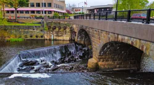 A stone bridge over a river with a small waterfall, surrounded by greenery and buildings in the background.