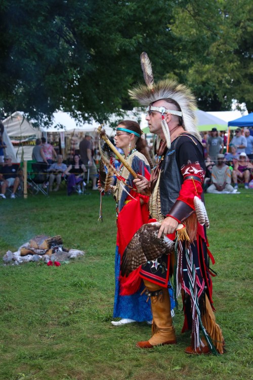 Two Indigenous men in traditional attire stand by a fire, surrounded by a gathering in a grassy area.