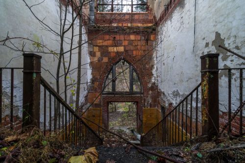 A view of a dilapidated building interior with crumbling walls, a staircase, and overgrown vegetation.