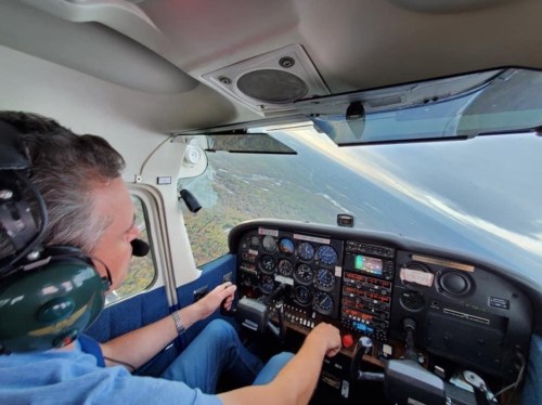 A pilot in a cockpit, focused on flying, with various instruments and a scenic view outside the window.