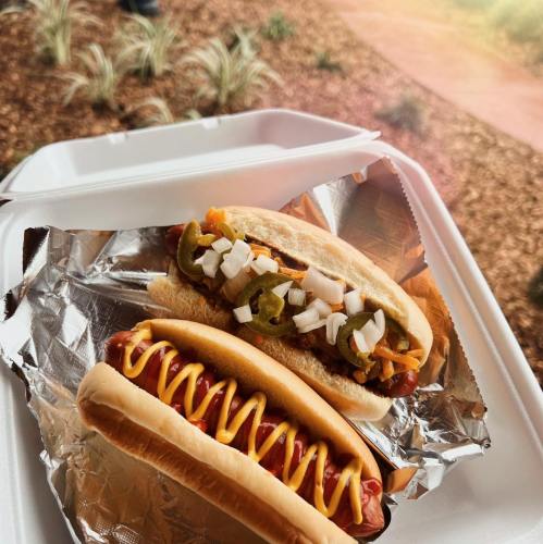 Two hot dogs in a takeout container, one topped with jalapeños and onions, the other with mustard.