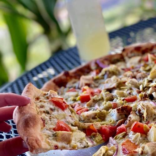 A hand reaches for a slice of pizza topped with vegetables, set on a table with a drink in the background.