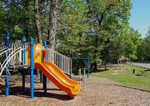 A playground with an orange slide surrounded by trees and grassy areas, featuring picnic tables in the background.