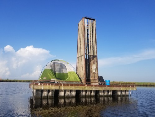 A green and gray tent on a wooden platform in a calm waterway, with a tall structure and blue sky in the background.