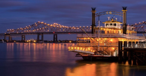 A brightly lit riverboat docked by a bridge at night, reflecting on the calm water.