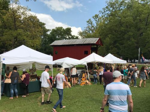A lively outdoor market with white tents, vendors, and visitors, set against a backdrop of trees and a red barn.