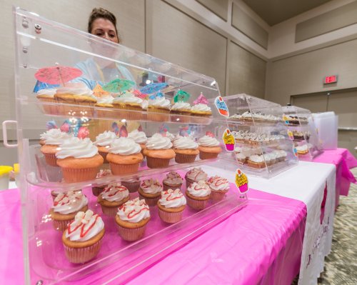 A display of colorful cupcakes in clear containers, with a person in the background arranging them.