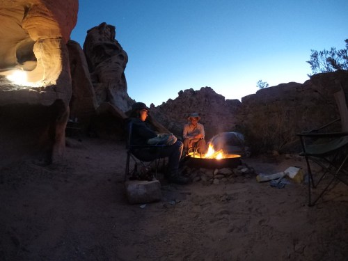 Two people sit by a campfire in a rocky desert landscape at dusk, surrounded by unique rock formations.