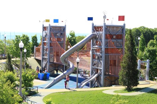 A modern playground with large slides, climbing structures, and colorful flags, surrounded by greenery and a lake view.