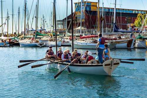 A group of people rowing a boat in a marina filled with sailboats and a colorful building in the background.