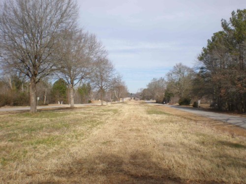 A wide, empty road lined with bare trees and grassy areas under a cloudy sky.