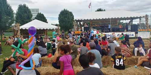 A crowd of children and adults sitting on hay bales at an outdoor event with a stage in the background.
