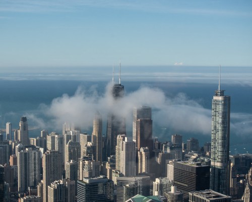 Aerial view of a city skyline with tall buildings partially shrouded in clouds, overlooking a body of water.