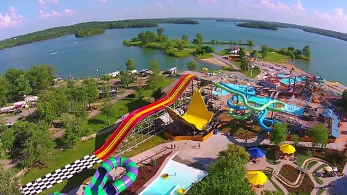 Aerial view of a water park with colorful slides, pools, and a lake in the background on a sunny day.