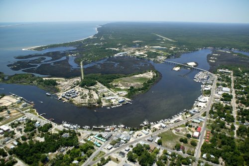 Aerial view of a coastal area featuring a river, marina, and surrounding greenery with a distant shoreline.