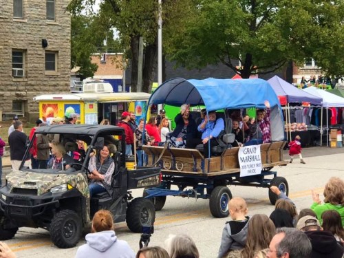 A parade scene featuring a decorated wagon with people waving, followed by a vehicle, amidst a crowd and vendor tents.