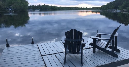 A serene lakeside view with two black chairs on a wooden dock, reflecting clouds and trees in the calm water.