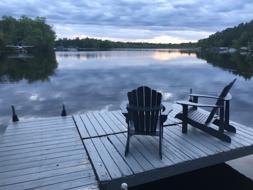 A serene lakeside dock with two chairs and a small table, reflecting a cloudy sky and calm water.