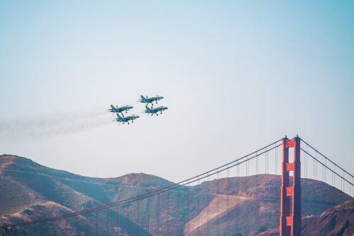 Four blue and yellow fighter jets flying in formation over the Golden Gate Bridge and hills in the background.