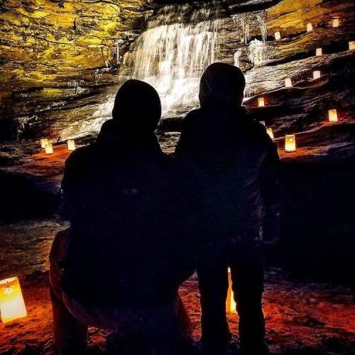 Two silhouetted figures sit in front of a waterfall, surrounded by glowing candles in a dark cave setting.