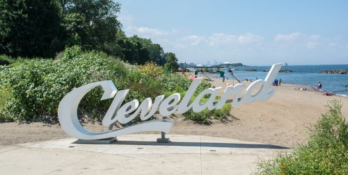 A large "Cleveland" sign on a sandy beach with people enjoying the sunny day and greenery in the background.