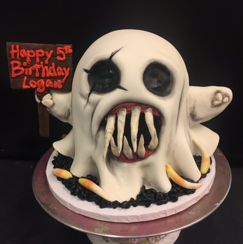 A spooky ghost-themed birthday cake with fangs, black details, and a sign that reads "Happy 5th Birthday Logan."